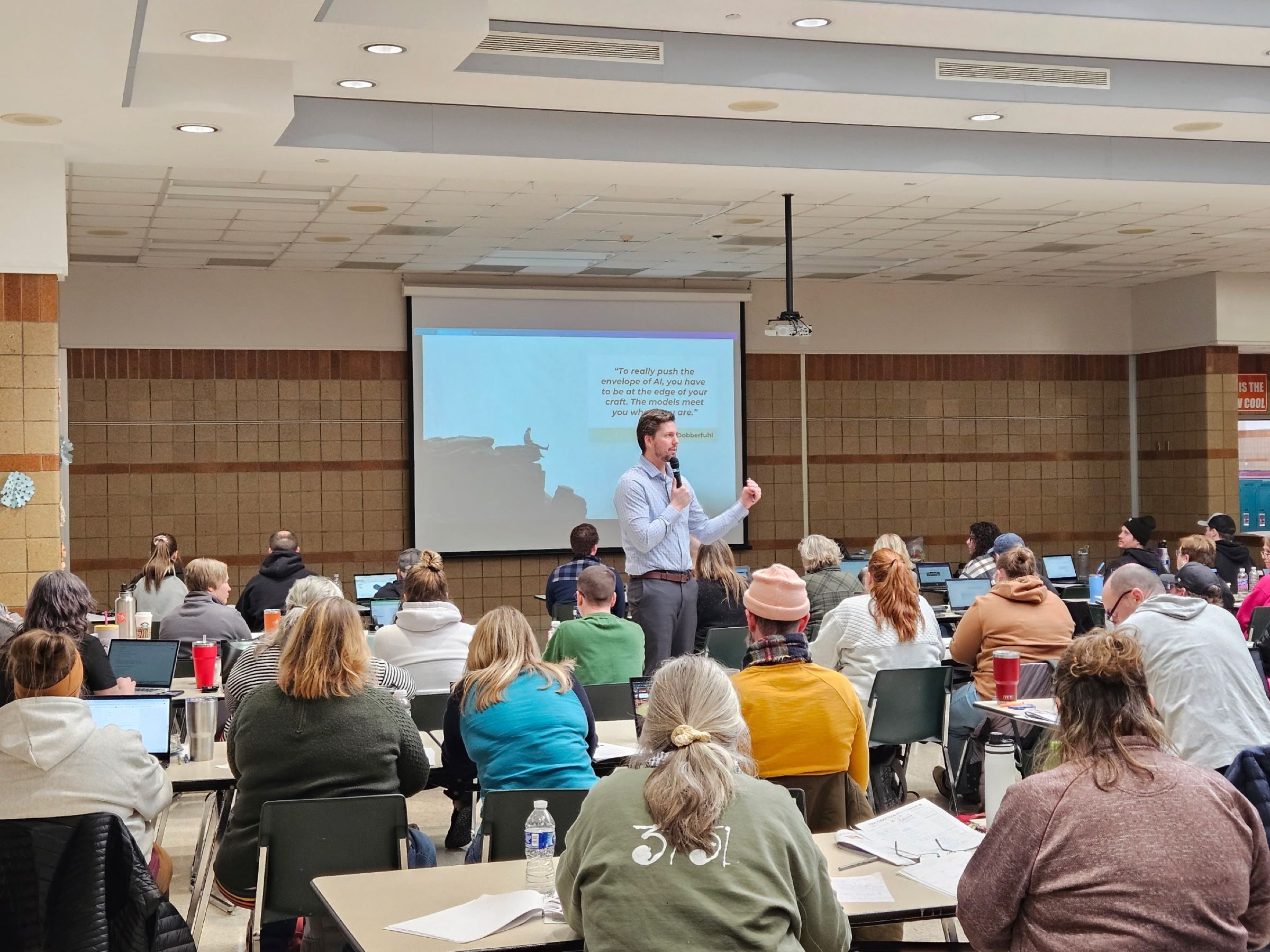 Tyler speaking with a microphone to a room of educators working on laptops in a school commons.